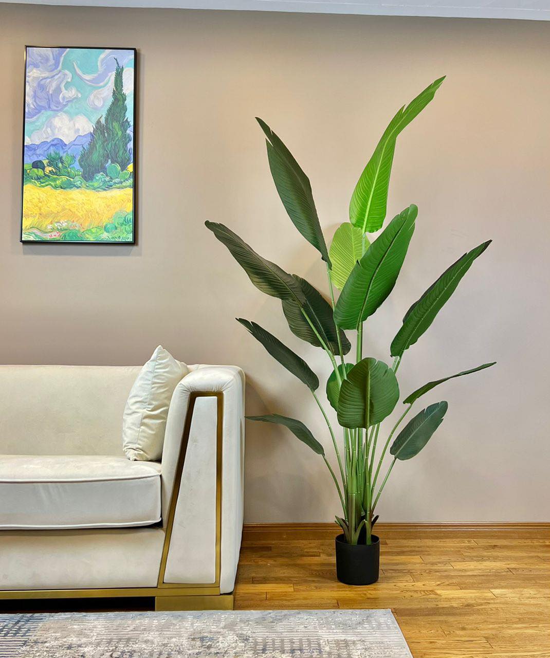 An elegant artificial bird of paradise plant with large green leaves is placed in a black planter on a hardwood floor beside a cream-colored modern sofa with gold accents. The room's beige walls create a warm backdrop, while a vibrant framed painting of a scenic landscape adds color and visual interest to the minimalist decor