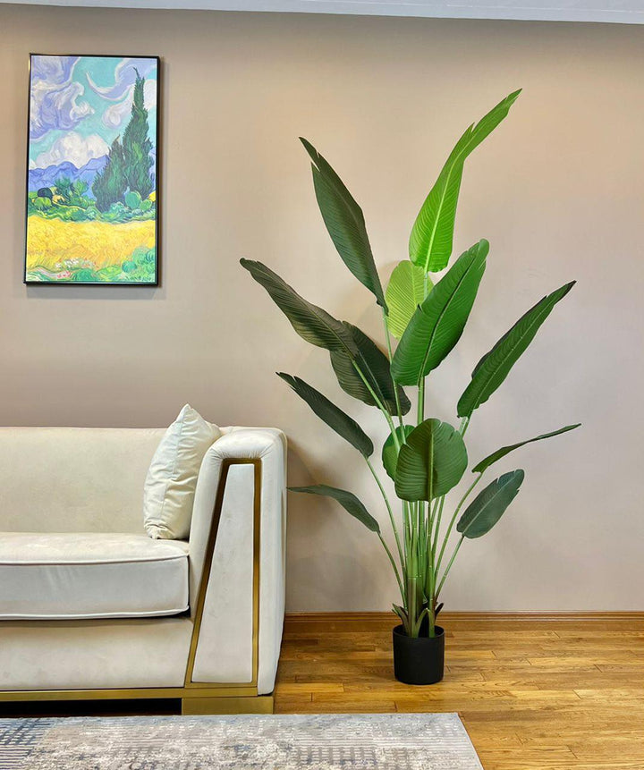An elegant artificial bird of paradise plant with large green leaves is placed in a black planter on a hardwood floor beside a cream-colored modern sofa with gold accents. The room's beige walls create a warm backdrop, while a vibrant framed painting of a scenic landscape adds color and visual interest to the minimalist decor