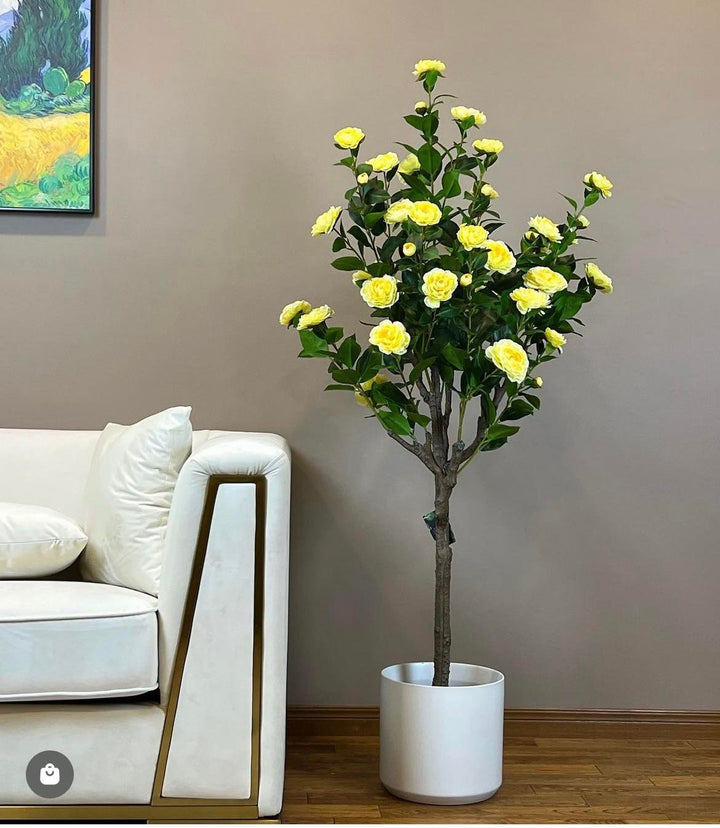 Artificial yellow flower tree in a white pot, placed next to a modern white sofa in a cozy living room setting, adding a bright and cheerful touch to the decor.