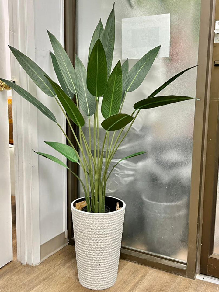 Artificial bird of paradise plant with multiple green leaves placed in a textured white planter, situated indoors next to a frosted glass door and a wooden floor