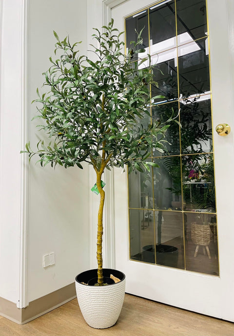 Artificial olive tree with slender green leaves and small black olives in a textured white pot placed near a glass-paneled door in a bright indoor space with wooden flooring.