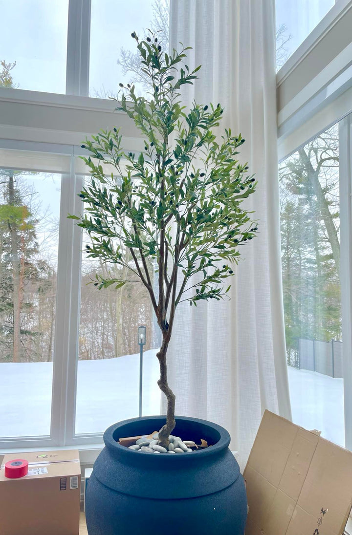 A tall artificial olive tree placed in a large, dark blue planter filled with white stones, set in front of floor-to-ceiling windows in a modern, bright room with snowy outdoor scenery visible through the windows