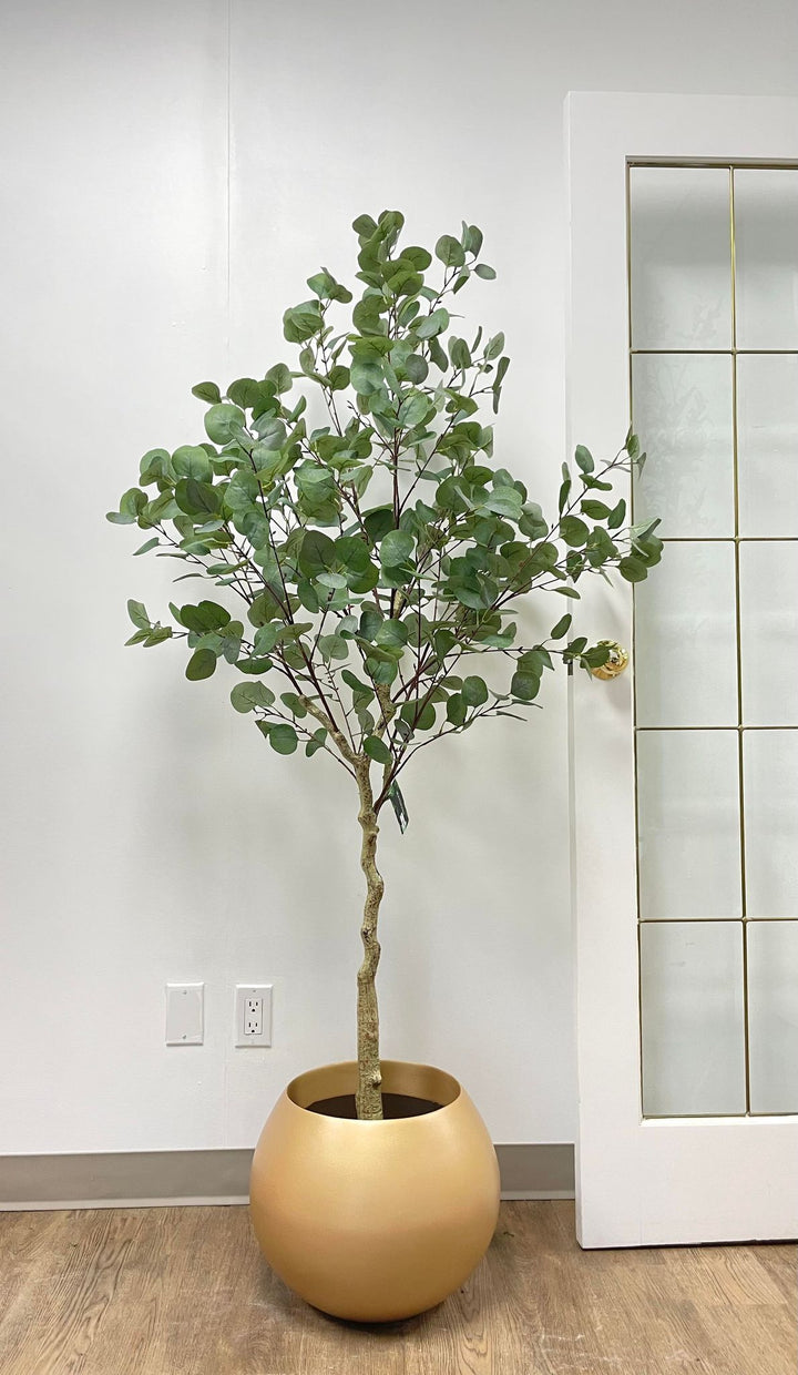 An artificial tree with lush green round leaves in a matte gold round planter, placed against a white wall next to a glass-paneled door in a minimalistic room.