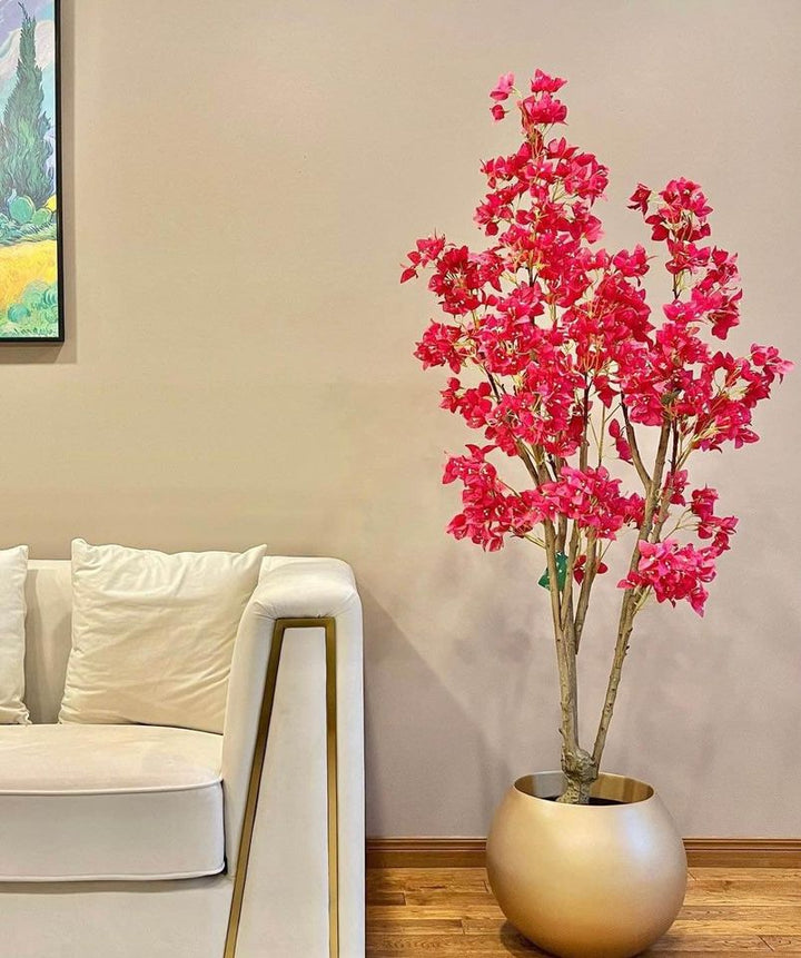 A bright artificial flowering plant with pink blossoms placed in a gold round planter next to a beige sofa, set against a neutral-toned wall in a modern living room.