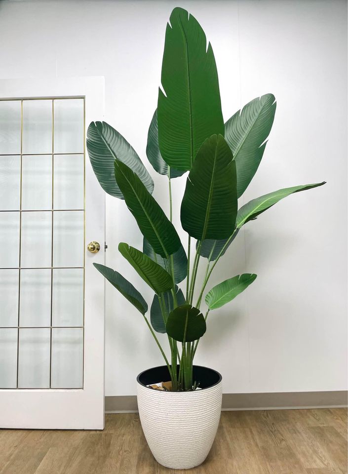 A large artificial bird of paradise plant in a white textured planter, placed on a light wood floor against a plain white wall. The plant features tall, broad green leaves that fan out gracefully, creating a lush and vibrant focal point in the room. A glass-paneled door with white trim is visible in the background, adding a touch of elegance to the minimalist interior setting.