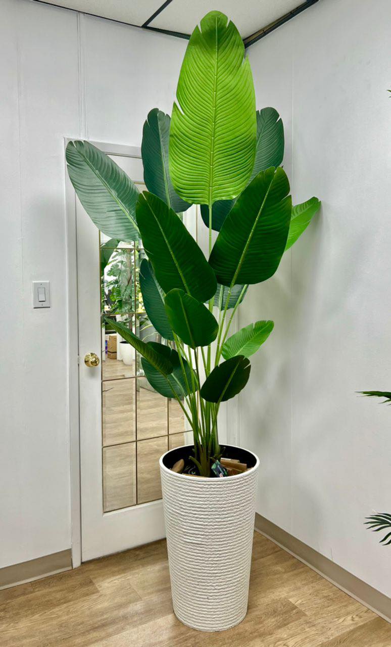 An elegant artificial bird of paradise plant with large green leaves is placed in a black planter on a hardwood floor beside a cream-colored modern sofa with gold accents. The room's beige walls create a warm backdrop, while a vibrant framed painting of a scenic landscape adds color and visual interest to the minimalist decor