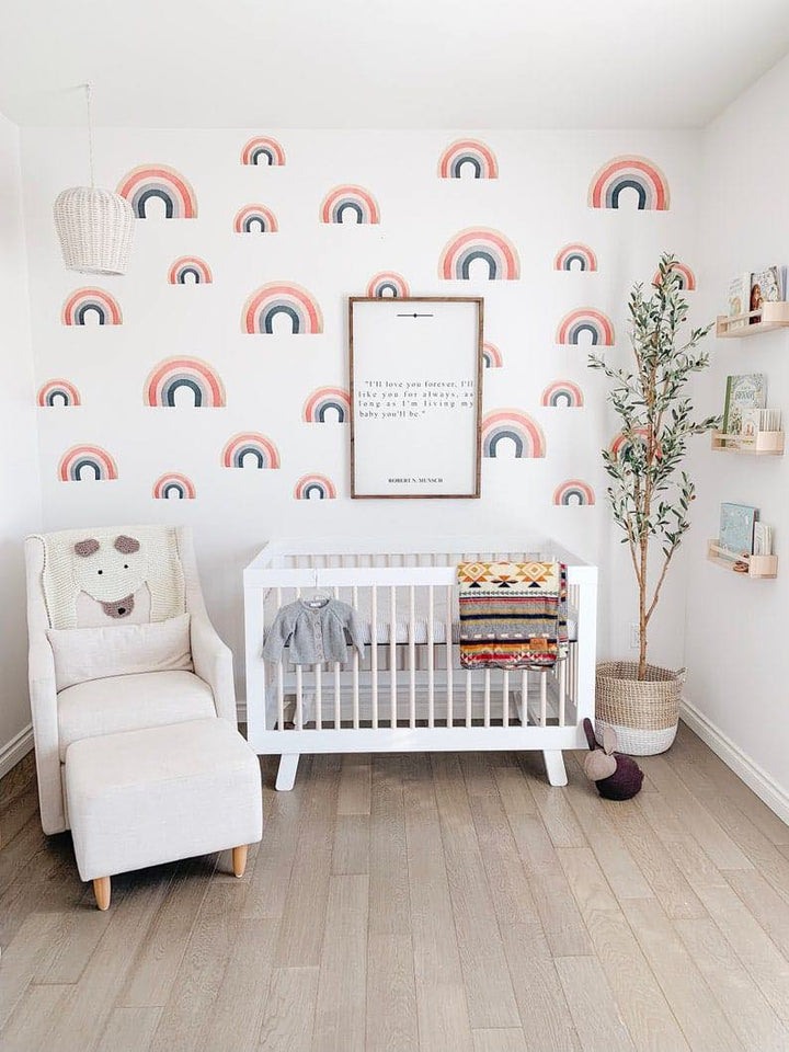 A cozy nursery with a rainbow-themed accent wall, featuring a white crib, a beige armchair with an ottoman, and an artificial olive tree in a woven basket.
