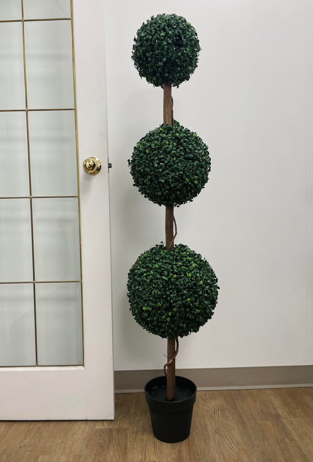 a three-tier artificial topiary tree in a black planter next to a beautiful door, perfect for enhancing indoor spaces with a touch of greenery and style