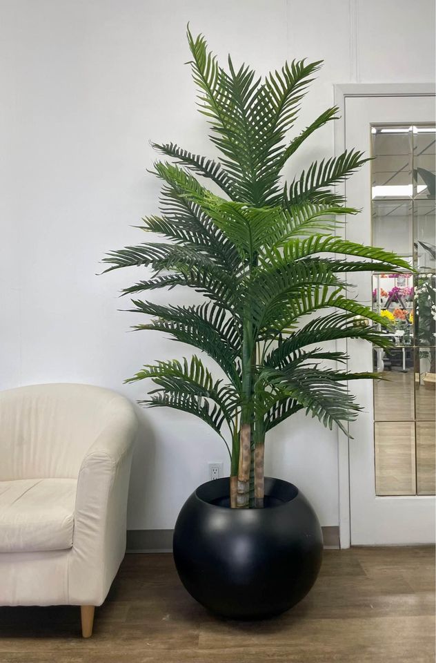 An indoor artificial palm plant placed in a large black round planter, next to a beige armchair in a modern room with wooden flooring and white walls.