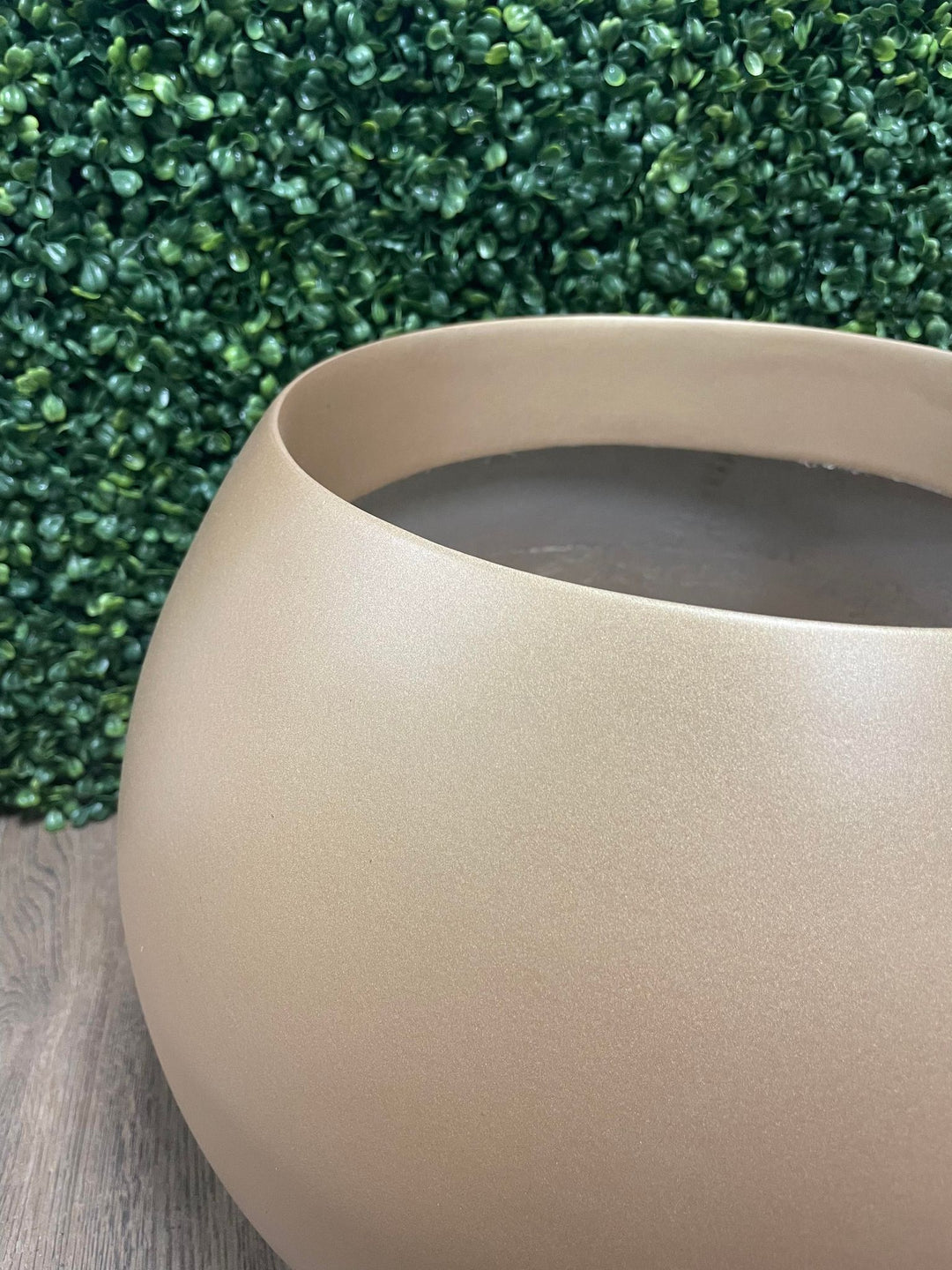 An artificial tree with lush green round leaves in a matte gold round planter, placed against a white wall next to a glass-paneled door in a minimalistic room.