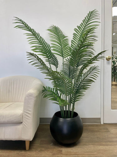 A tall, artificial palm plant is placed in a matte black, round planter. The plant's lush green leaves fan out elegantly, complementing the minimalist indoor space. To the left is a soft beige armchair, and in the background is a white wall with a glass-paneled door, adding to the modern, clean aesthetic of the room.