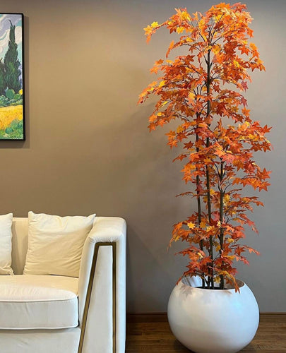 A bright orange artificial tree in a white spherical planter placed next to a cream-colored sofa against a beige wall, with a painting hung on the left side.