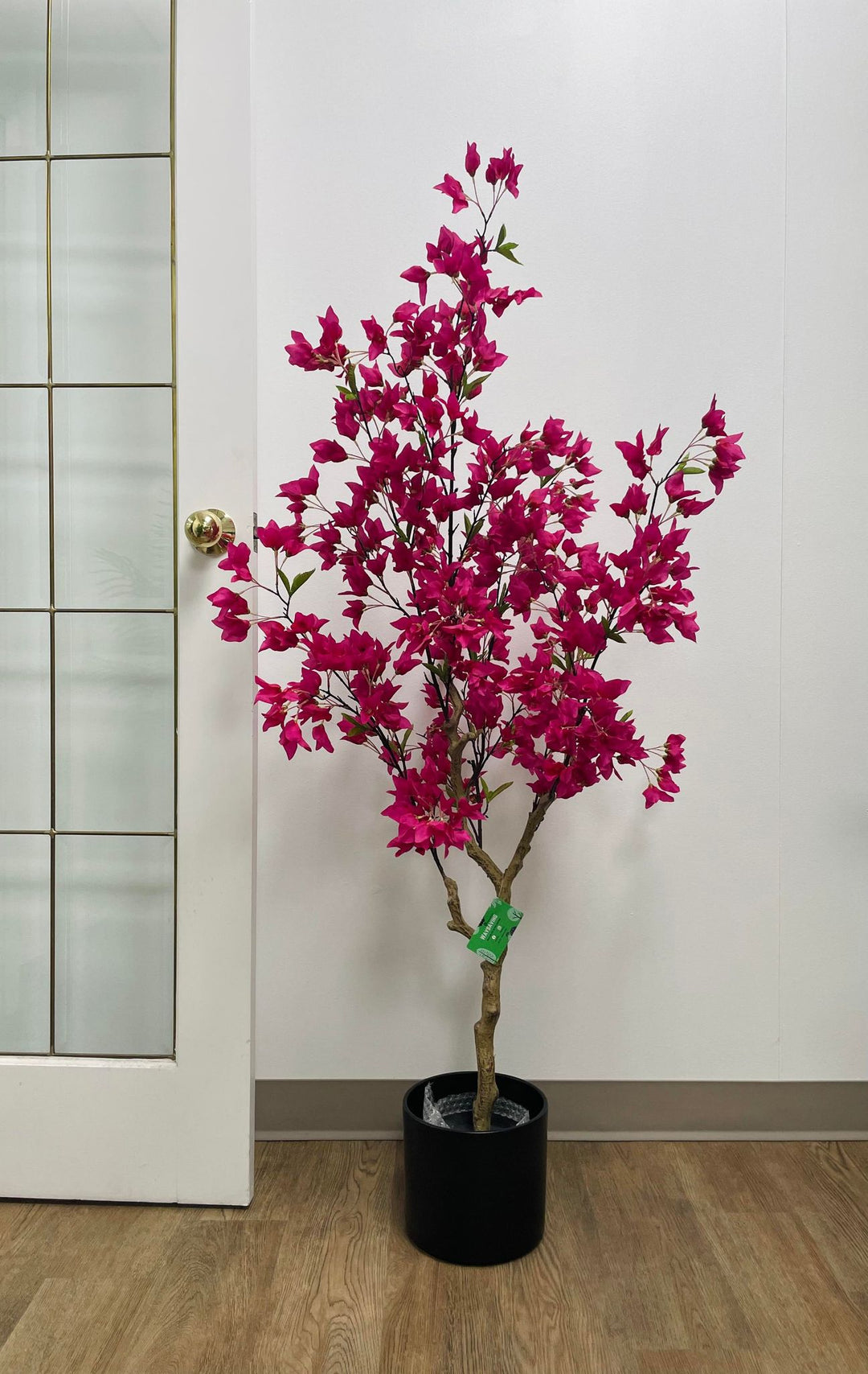 Artificial pink flowering tree in a black planter, placed on wooden flooring next to a glass-paneled door in a modern indoor setting