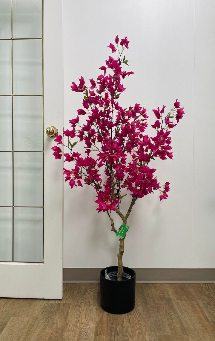 Artificial pink flowering tree in a black planter, placed on wooden flooring next to a glass-paneled door in a modern indoor setting