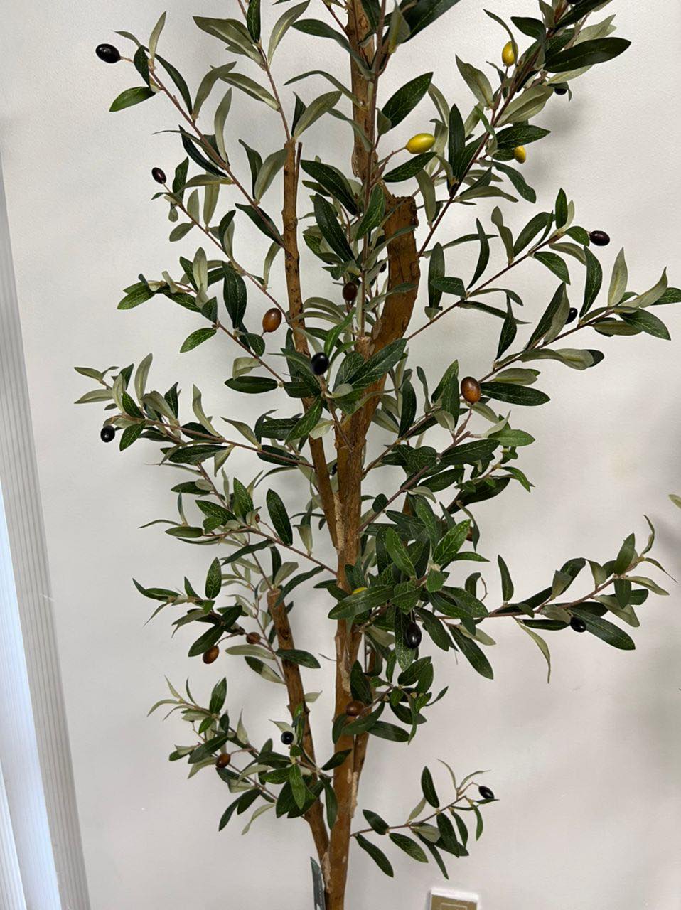 Artificial olive tree with dense green foliage and small black olives, standing against a neutral-colored wall. The leaves have a natural texture and veining, creating a realistic appearance.