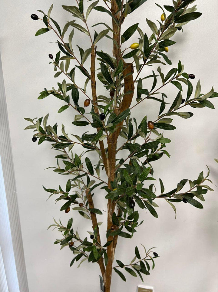 Artificial olive tree with dense green foliage and small black olives, standing against a neutral-colored wall. The leaves have a natural texture and veining, creating a realistic appearance.