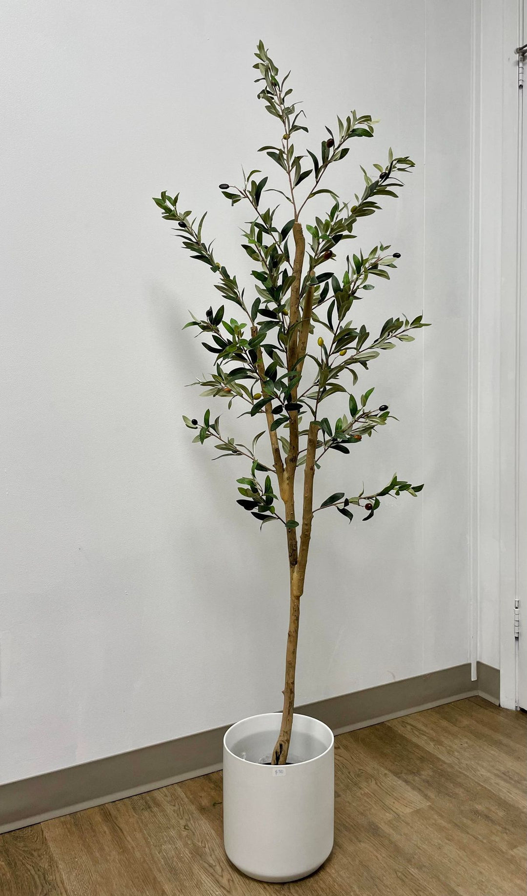 Artificial olive tree in a sleek white pot, placed on a wooden floor against a plain white wall.