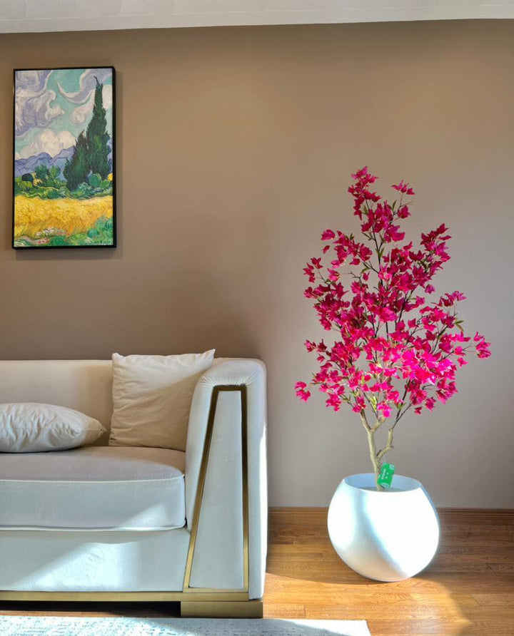 Artificial pink flowering tree in a white round planter, placed next to a beige sofa in a modern living room with wooden flooring.
