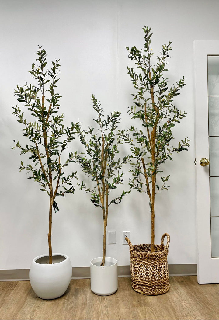 Three artificial olive trees of varying heights are displayed indoors, each in a different container—one in a round white pot, one in a cylindrical white pot, and the third in a woven basket