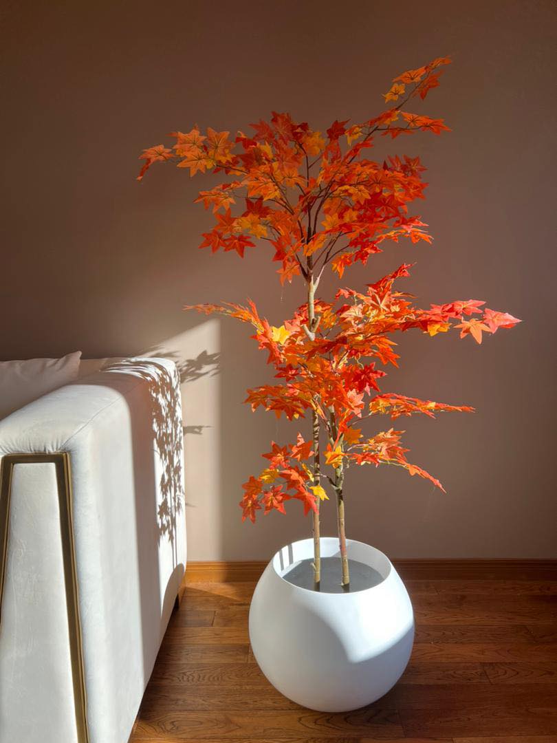Artificial autumn maple tree with vibrant orange and red leaves in a white round pot, placed next to a modern white sofa against a warm-toned wall, with sunlight casting shadows on the floor