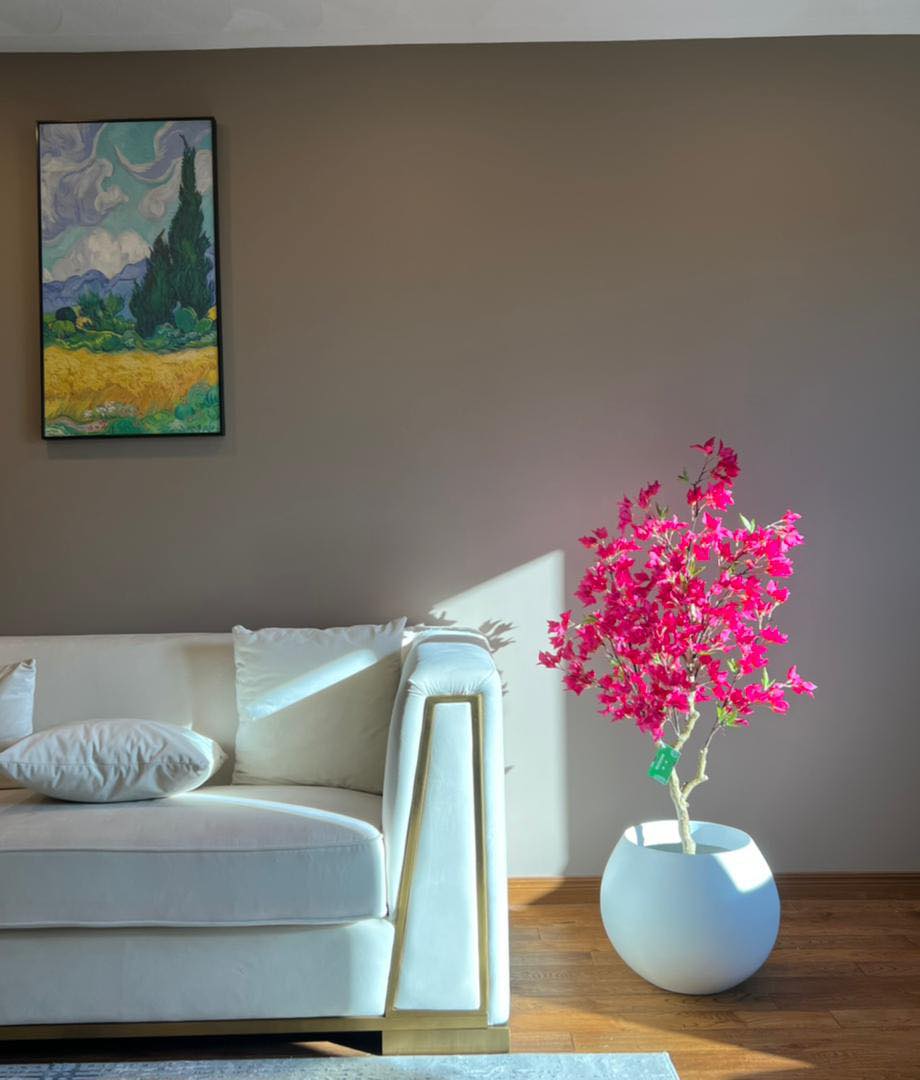 Artificial pink flowering tree in a white pot, placed beside a cream-colored couch with sunlight streaming in, highlighting the vibrant colors in a cozy living room setting
