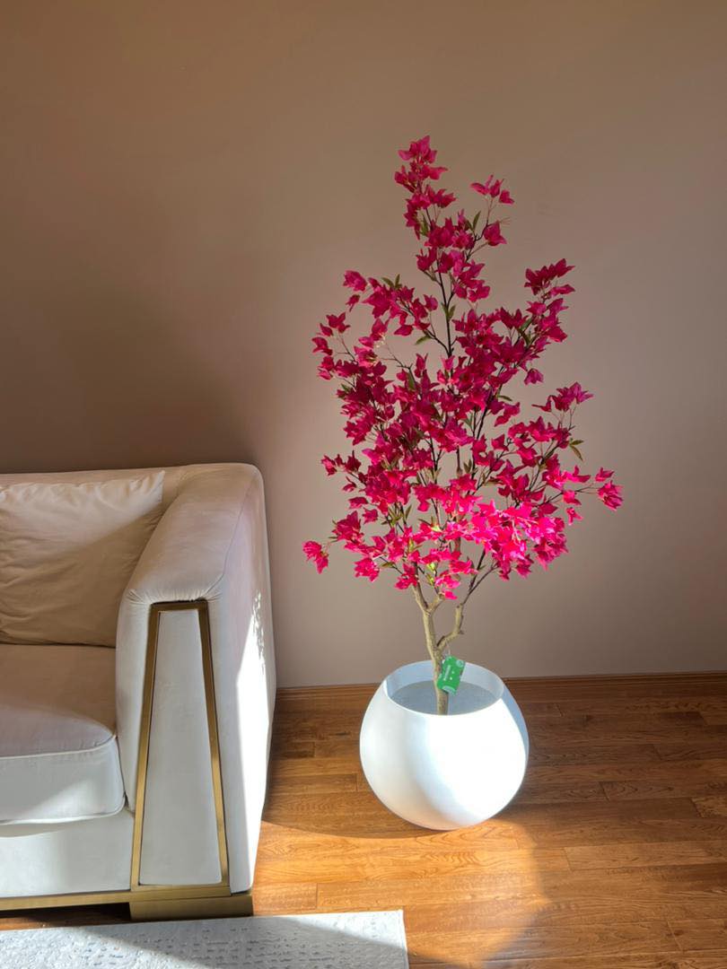 Artificial pink flowering tree in a white round planter, placed next to a beige sofa in a modern living room with wooden flooring.