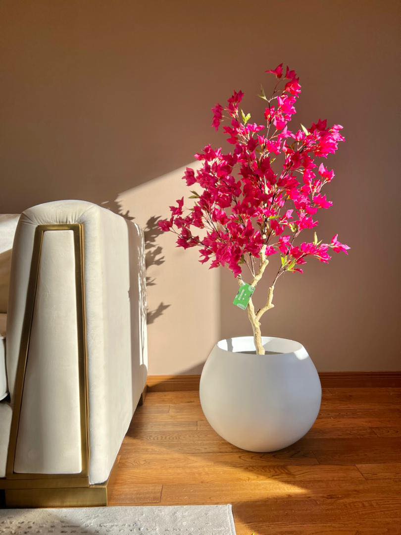 Artificial pink flowering tree in a white pot placed next to a modern beige sofa, with sunlight casting shadows on a wooden floor