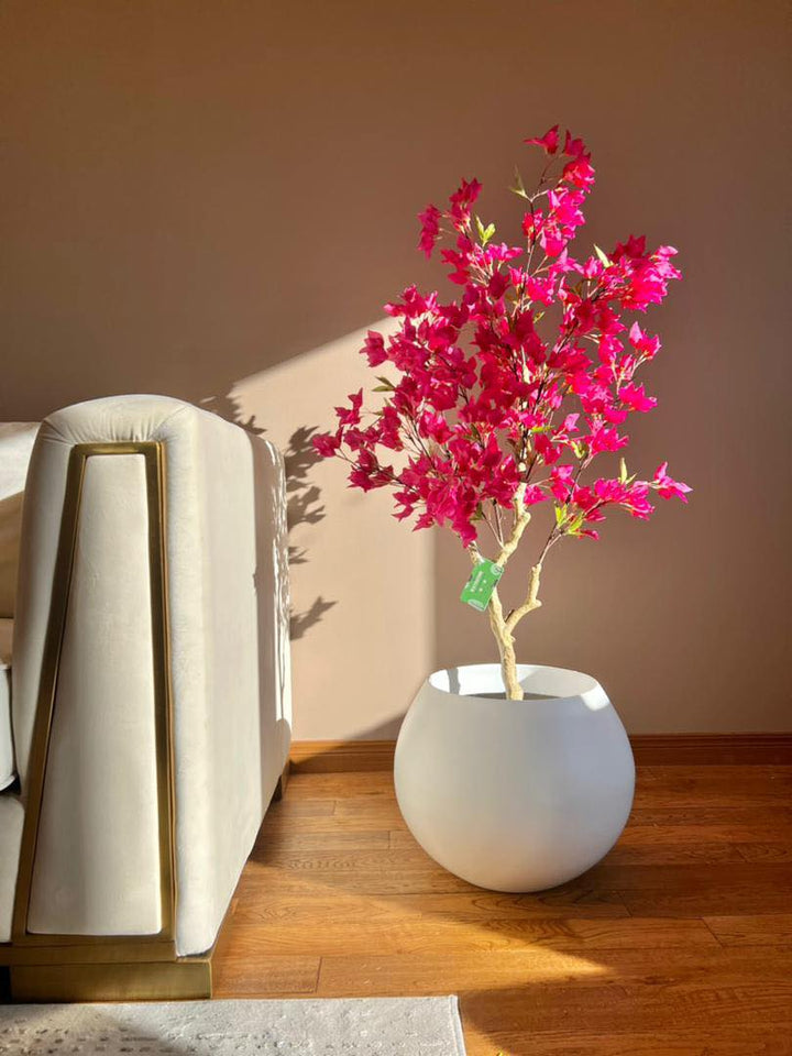 Artificial pink flowering tree in a white pot placed next to a modern beige sofa, with sunlight casting shadows on a wooden floor
