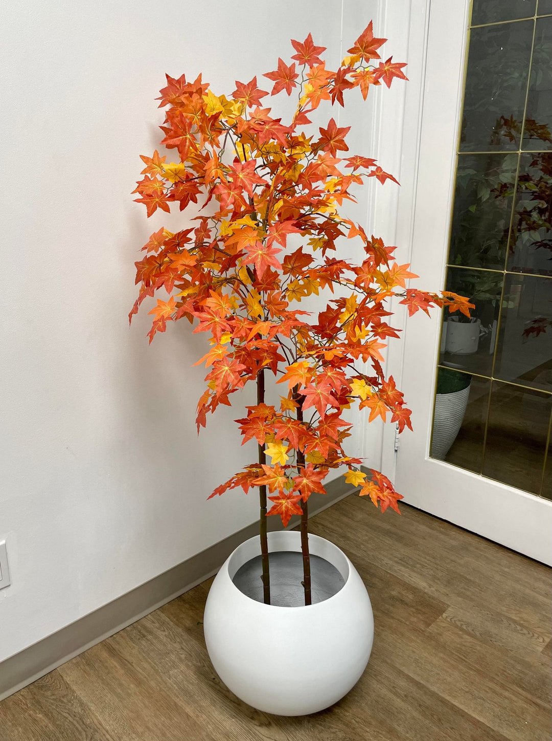 Artificial autumn maple tree with vibrant orange and red leaves in a white round pot, placed next to a modern white sofa against a warm-toned wall, with sunlight casting shadows on the floor