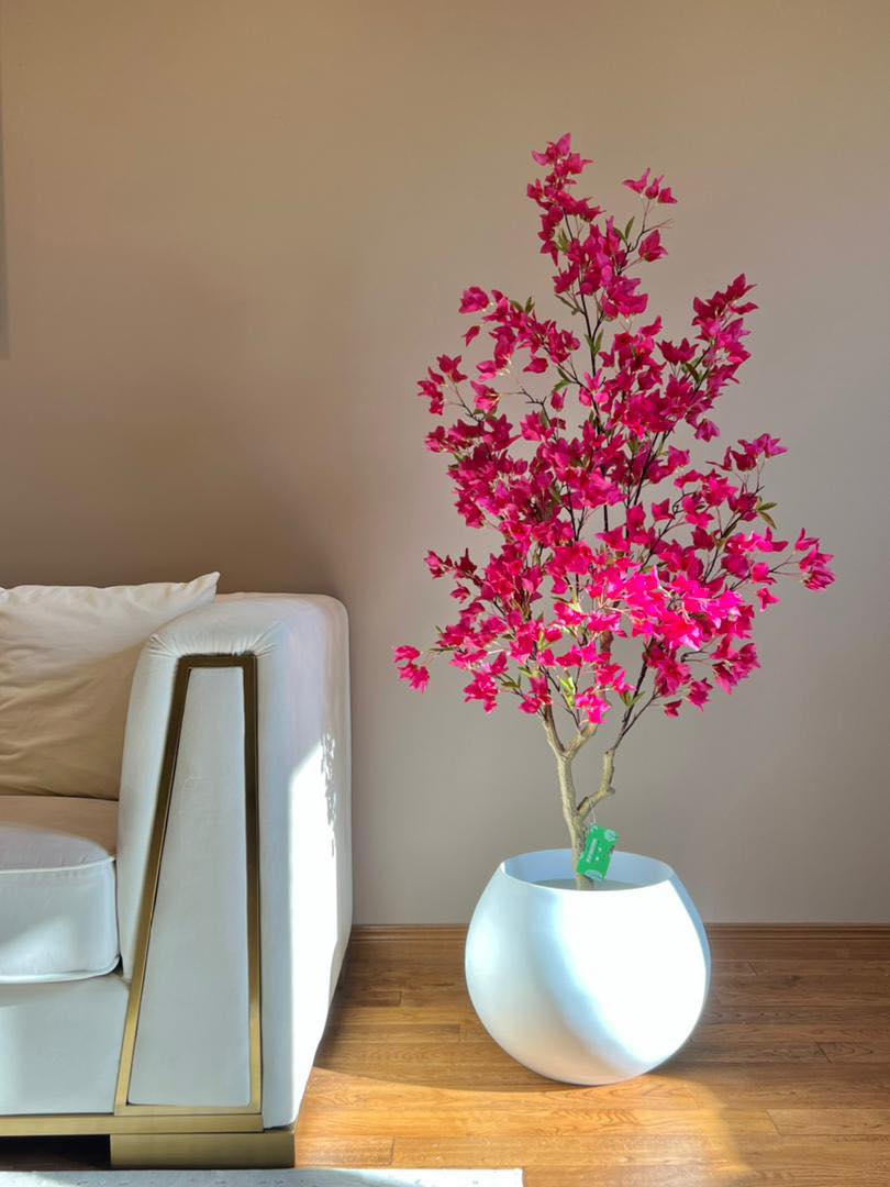 Artificial pink flowering tree in a white round planter, placed next to a beige sofa in a modern living room with wooden flooring