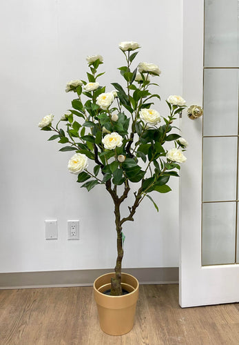 This is an artificial flowering tree with white blossoms, placed in a simple tan pot. The tree has several branches with lush green leaves and multiple white flowers, creating a natural and elegant appearance. It's positioned indoors against a white wall, near a glass door. The overall look is minimalistic and clean, making it suitable for both home and office decor