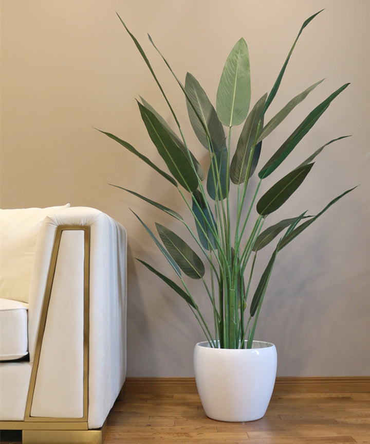 A sleek and tall artificial bird of paradise plant with elongated green leaves is placed in a modern, white round pot. The plant stands beside a contemporary white couch with gold accents, adding a touch of nature to the minimalist and sophisticated living room decor. The warm wood floor complements the overall neutral and elegant color scheme.