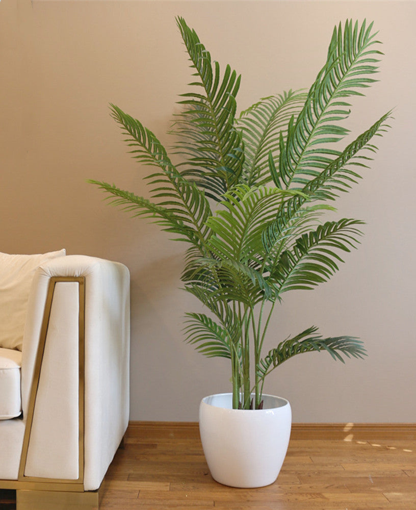 A tall artificial palm tree with vibrant green leaves, placed in a simple black pot on a wooden floor, next to a modern white couch and a neutral-toned wall. The tree adds a lush, tropical feel to the space.