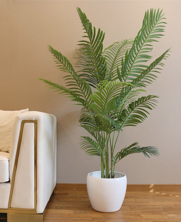 A tall artificial palm tree with vibrant green leaves, placed in a simple black pot on a wooden floor, next to a modern white couch and a neutral-toned wall. The tree adds a lush, tropical feel to the space.