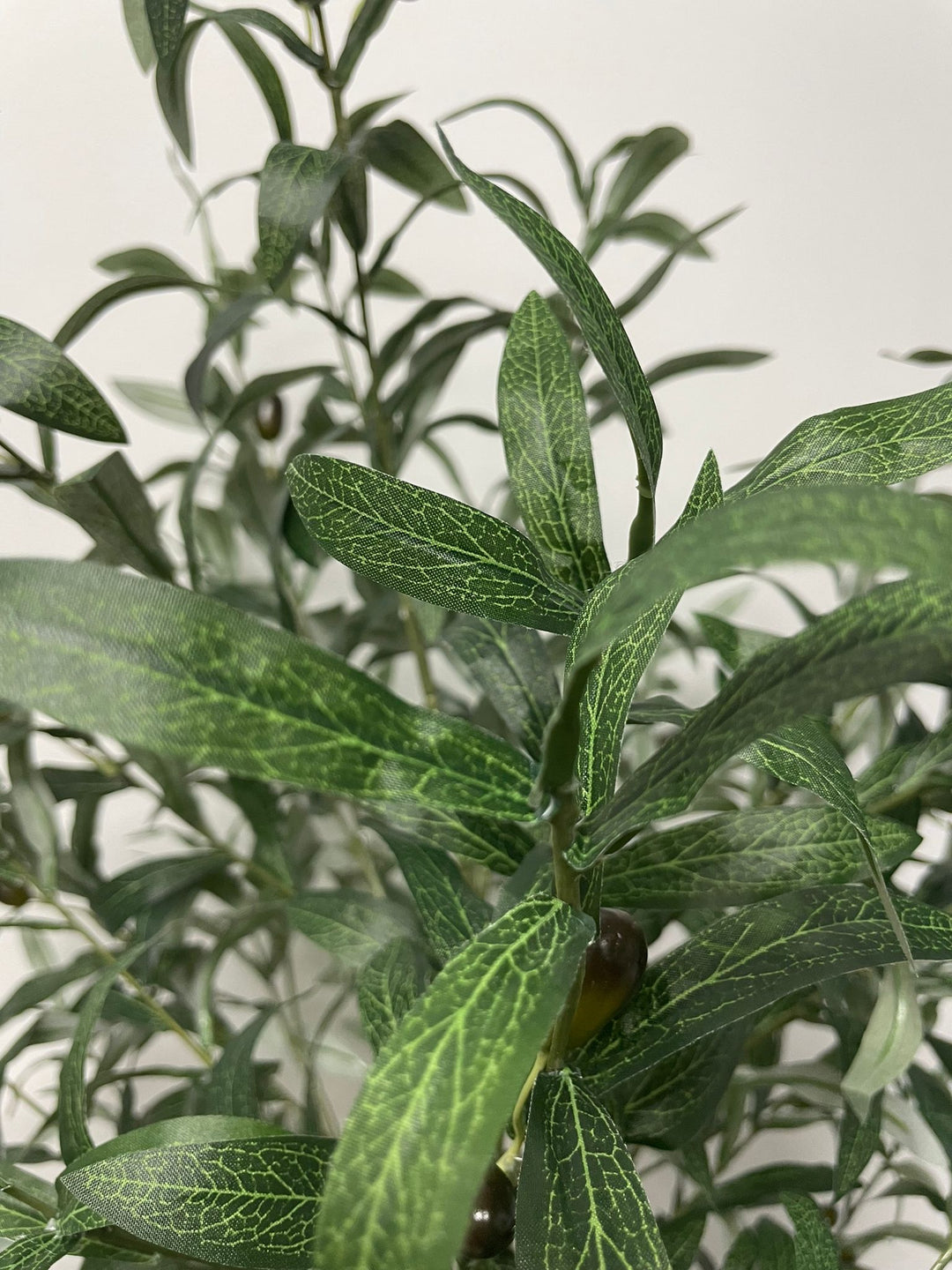 Close-up of artificial olive tree leaves, showcasing the detailed veining and texture of the green foliage, with a few black olives visible in the background.