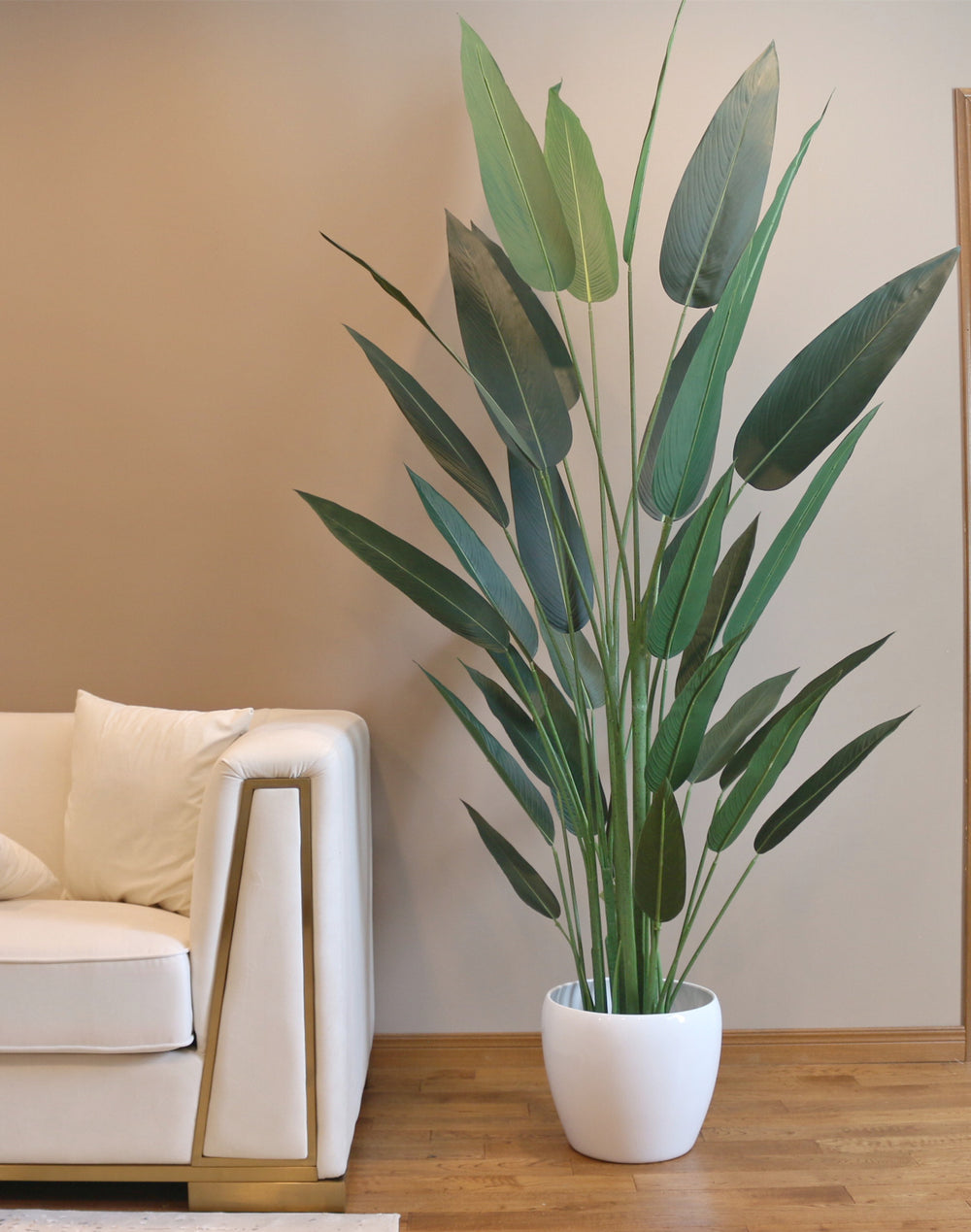 Large 7ft artificial bird of paradise plant with elongated green leaves, placed in a white planter next to a cream-colored couch in a modern living room.