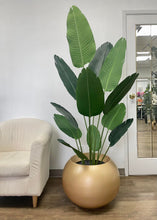 Charger l&#39;image dans la galerie, Large artificial tropical plant with broad, dark green leaves placed in a matte gold spherical planter, next to a cream-colored armchair on a wooden floor.
