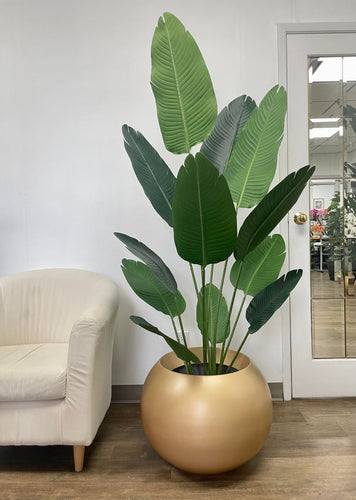 Large artificial tropical plant with broad, dark green leaves placed in a matte gold spherical planter, next to a cream-colored armchair on a wooden floor.