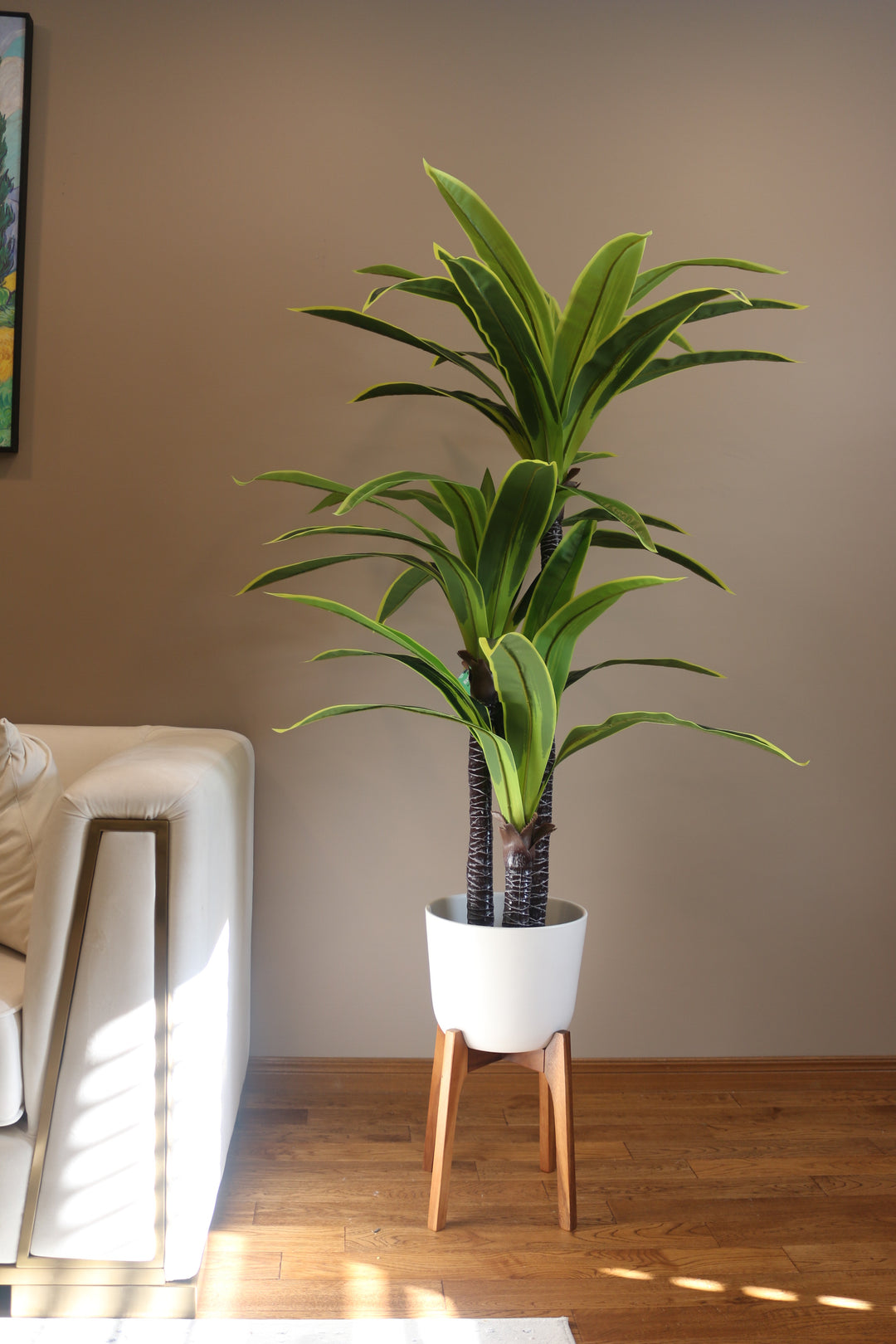 Artificial tall green plant with broad, vibrant leaves in a sleek white pot, paired with a modern cream sofa, perfect for contemporary home or office decor
