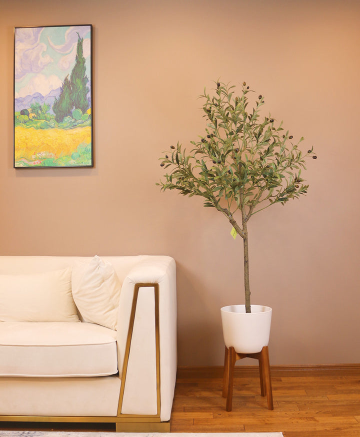 Elegant living room setup featuring a tall artificial olive tree in a white pot positioned next to a modern beige sofa with gold trim. The room has a warm, brown-toned wall adorned with a colorful landscape painting, creating a cozy and sophisticated atmosphere.