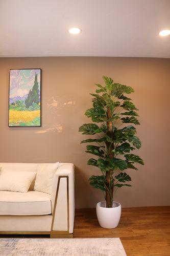 Artificial monstera plant in a white pot placed beside a modern beige sofa in a stylish living room. The lush green foliage adds a vibrant touch to the neutral-colored space, complemented by a colorful wall painting above the sofa, creating a cozy and inviting atmosphere