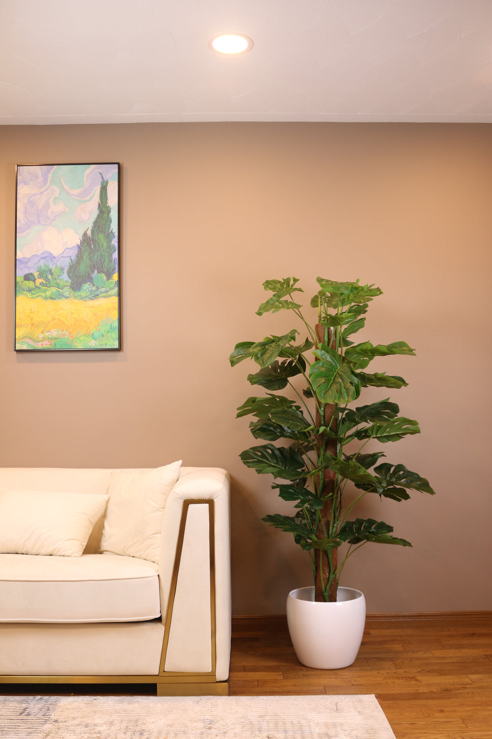 Artificial monstera plant in a white pot placed beside a modern beige sofa in a stylish living room. The lush green foliage adds a vibrant touch to the neutral-colored space, complemented by a colorful wall painting above the sofa, creating a cozy and inviting atmosphere