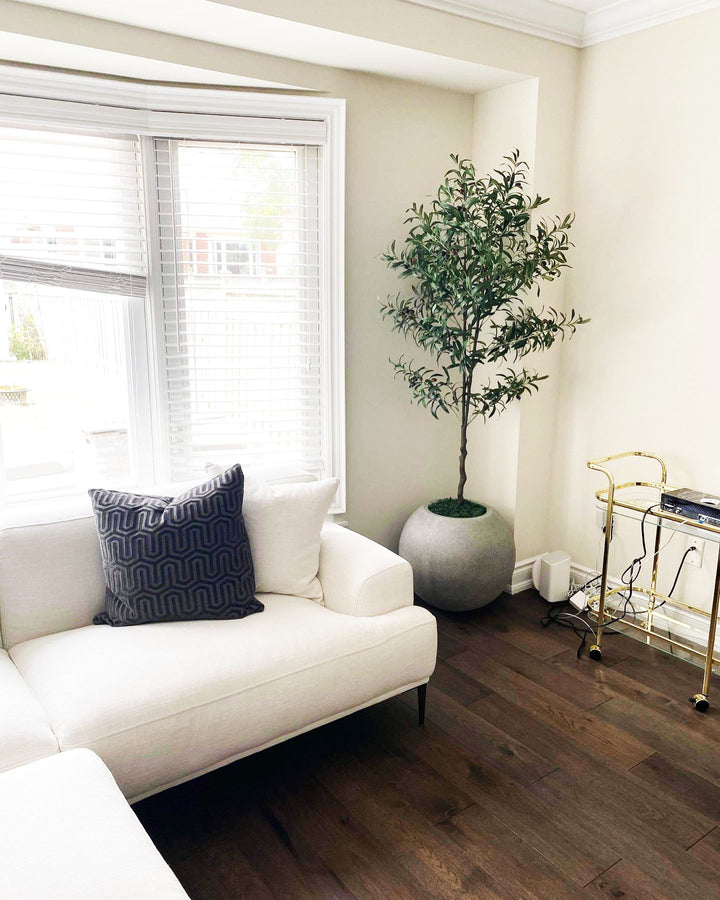  A cozy living room corner featuring a white sectional sofa with a dark decorative pillow, positioned near a large window with white blinds. To the right of the sofa, a realistic artificial olive tree is placed in a round gray planter on dark hardwood flooring. The corner is accented by a gold-colored bar cart, adding a touch of elegance to the modern, minimalist space. Natural light filters through the window, enhancing the warm and inviting ambiance of the room.