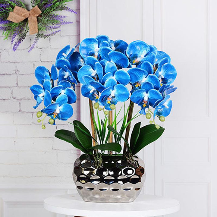 A vibrant arrangement of artificial blue orchids placed in a modern, metallic vase with a textured surface. The blue flowers pop against the light background of a white brick wall, and there is a small wreath hanging in the corner with lavender and a rustic bow for added decorative flair.