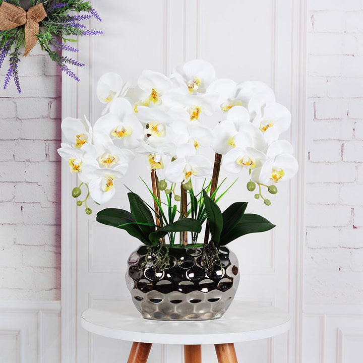 A stylish arrangement of artificial white orchids with yellow centers placed in a reflective silver vase. The vase has a distinct dimpled pattern and is positioned on a white round table with wooden legs. The background features a brick wall partially adorned with lavender and a rustic bow, creating a charming, cozy interior scene.