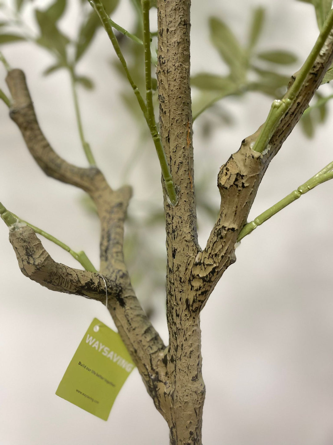 Close-up of the detailed trunk and branches of an artificial olive tree with a textured, lifelike appearance. A green tag with the brand name "Waysaving" is attached to one of the branches. The tree features realistic bark textures and connected branches, making it a convincing decorative piece for indoor settings.