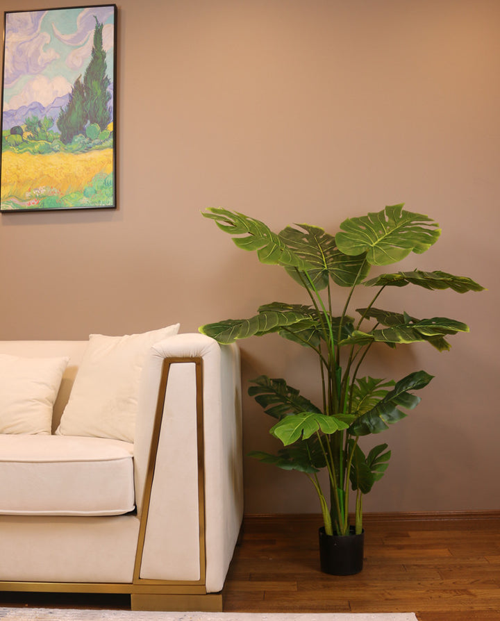 Artificial monstera plant in a black pot, placed on a wooden floor next to a modern white and gold-trimmed armchair. The plant features large, glossy green leaves with distinctive splits, creating a tropical atmosphere in the indoor setting