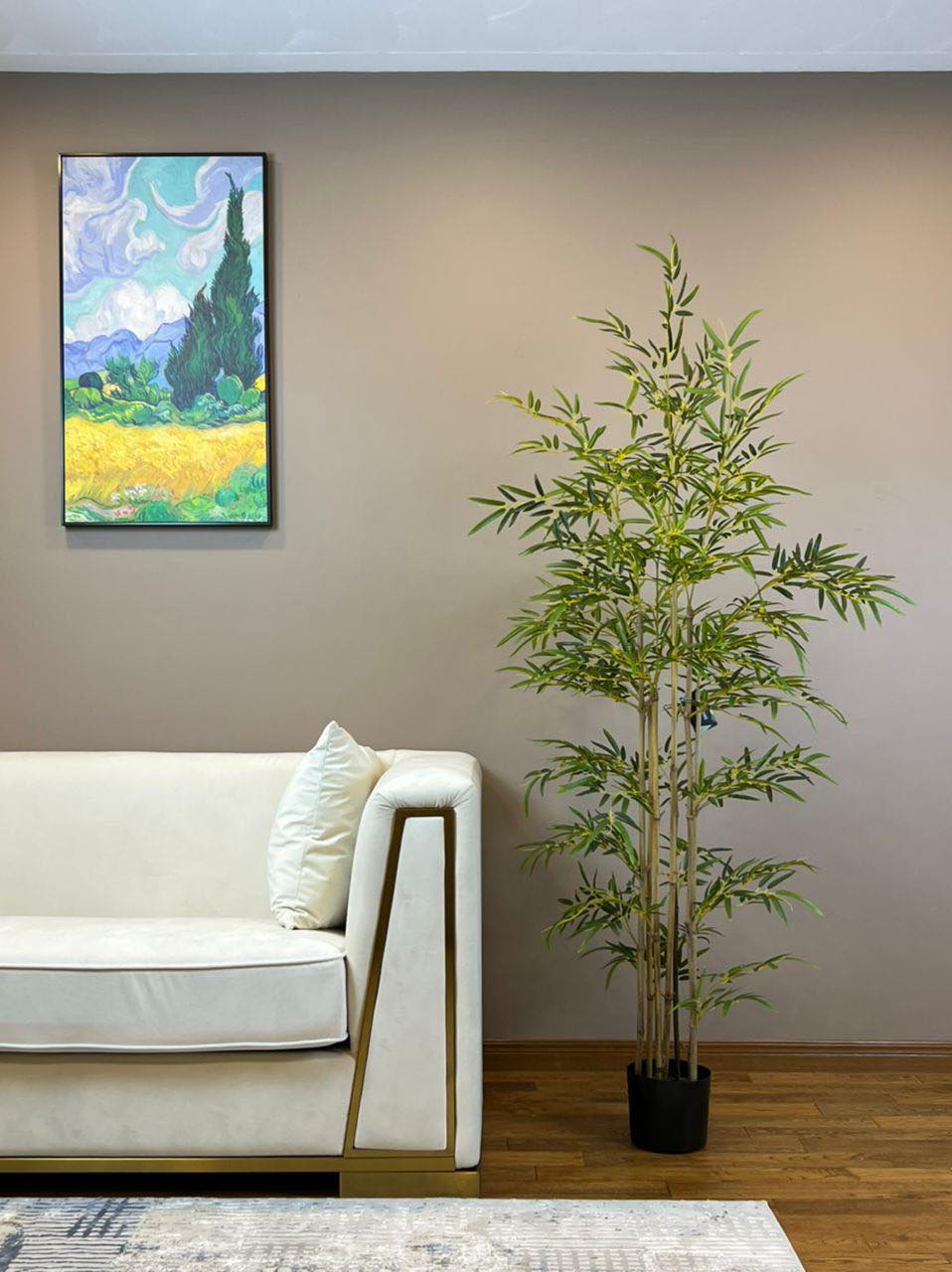 Artificial bamboo plant in a black nursery pot, placed beside a modern white sofa in a living room.