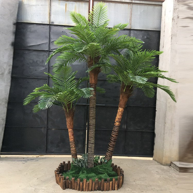 Artificial palm trees with coconuts set up indoors, surrounded by lush greenery and enclosed in a wooden planter, creating a vibrant tropical display perfect for brightening up any indoor space.