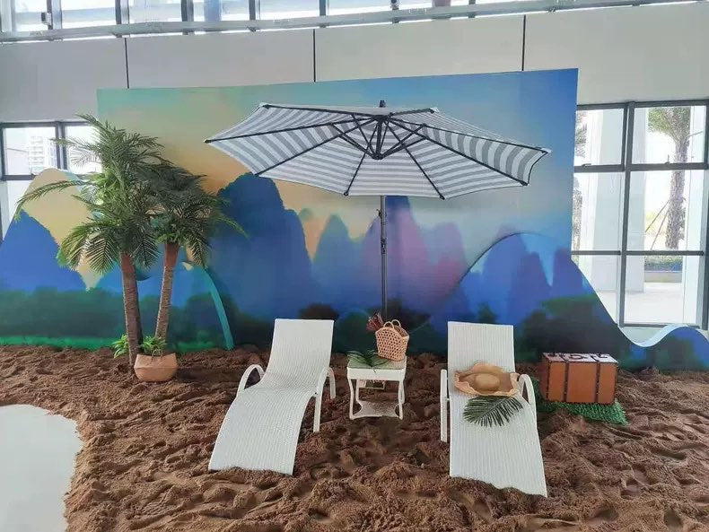 An indoor beach setup with two white lounge chairs, a large striped umbrella, and a small table. The area is decorated with artificial coconut trees  for a beachy photoshoot.